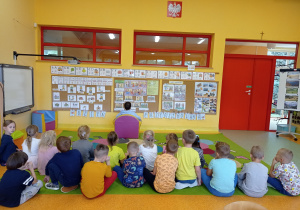 dzieci siedzą na dywanie w dwóch rzędach jedna dziecko siedzi tyłem do nich na pufce